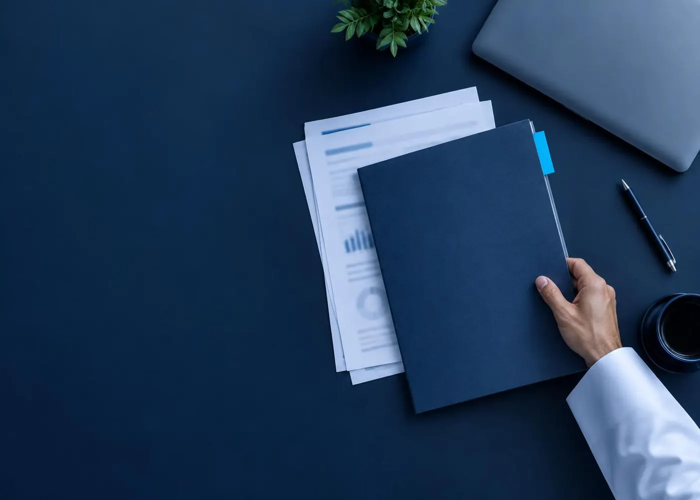 Top-down view of a desk with printed documents, a closed laptop and a hand placing a handover folder